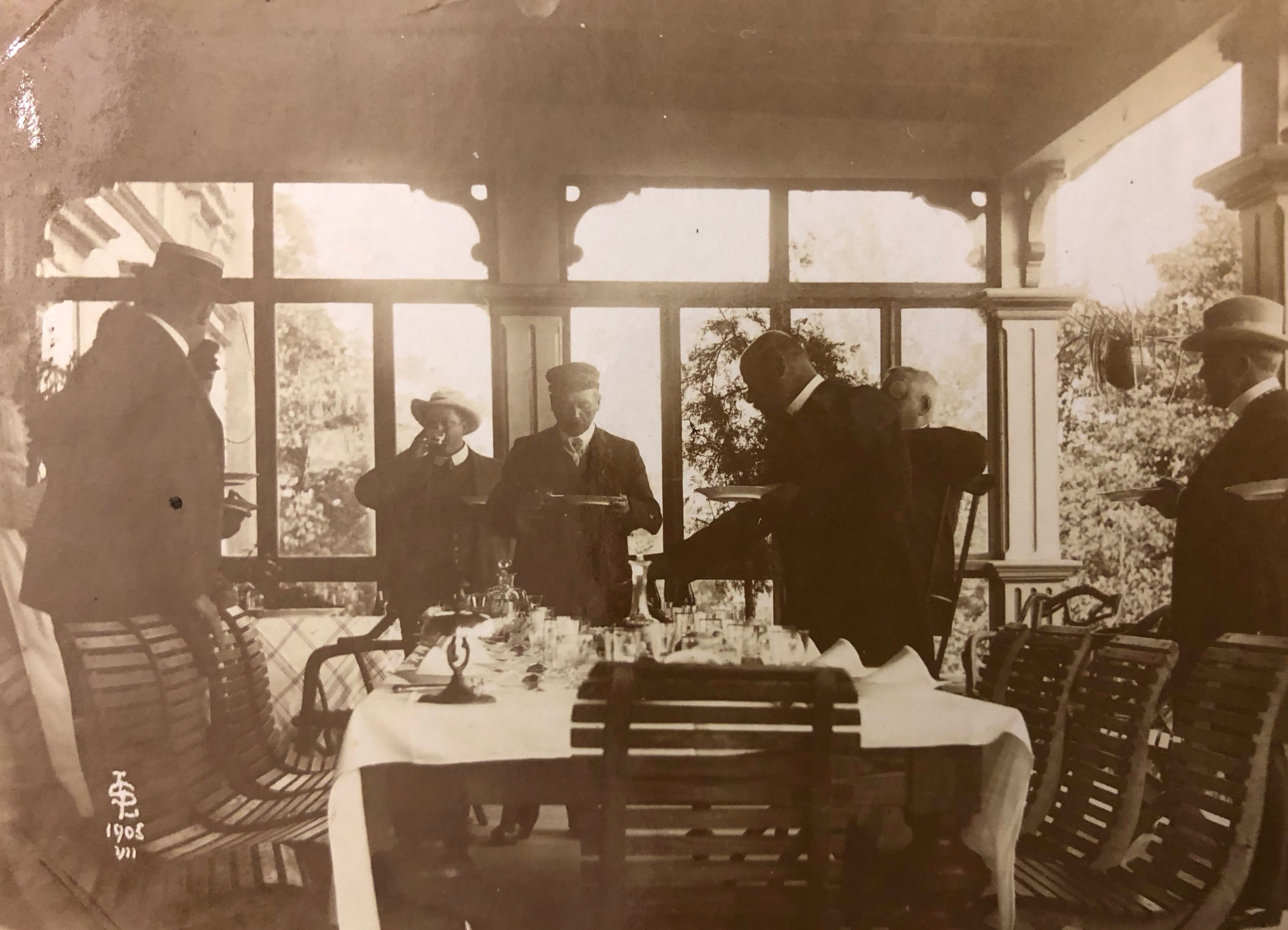 Svartvitt fotografi av ett sällskap festklädda herrar som står eller sitter runt ett stort bord på en inglasad veranda. 