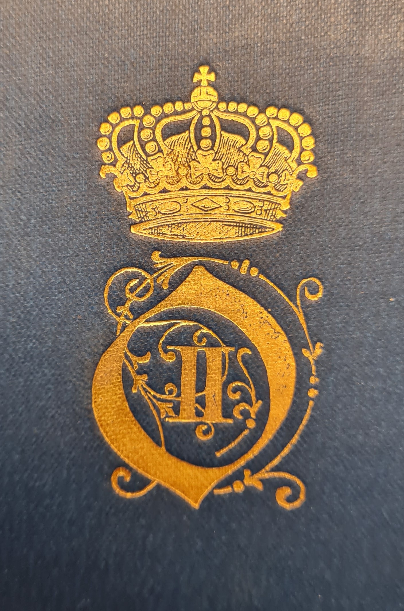 Färgfotografi av ett förgyllt monogram med kungakrona på en blå bokpärm