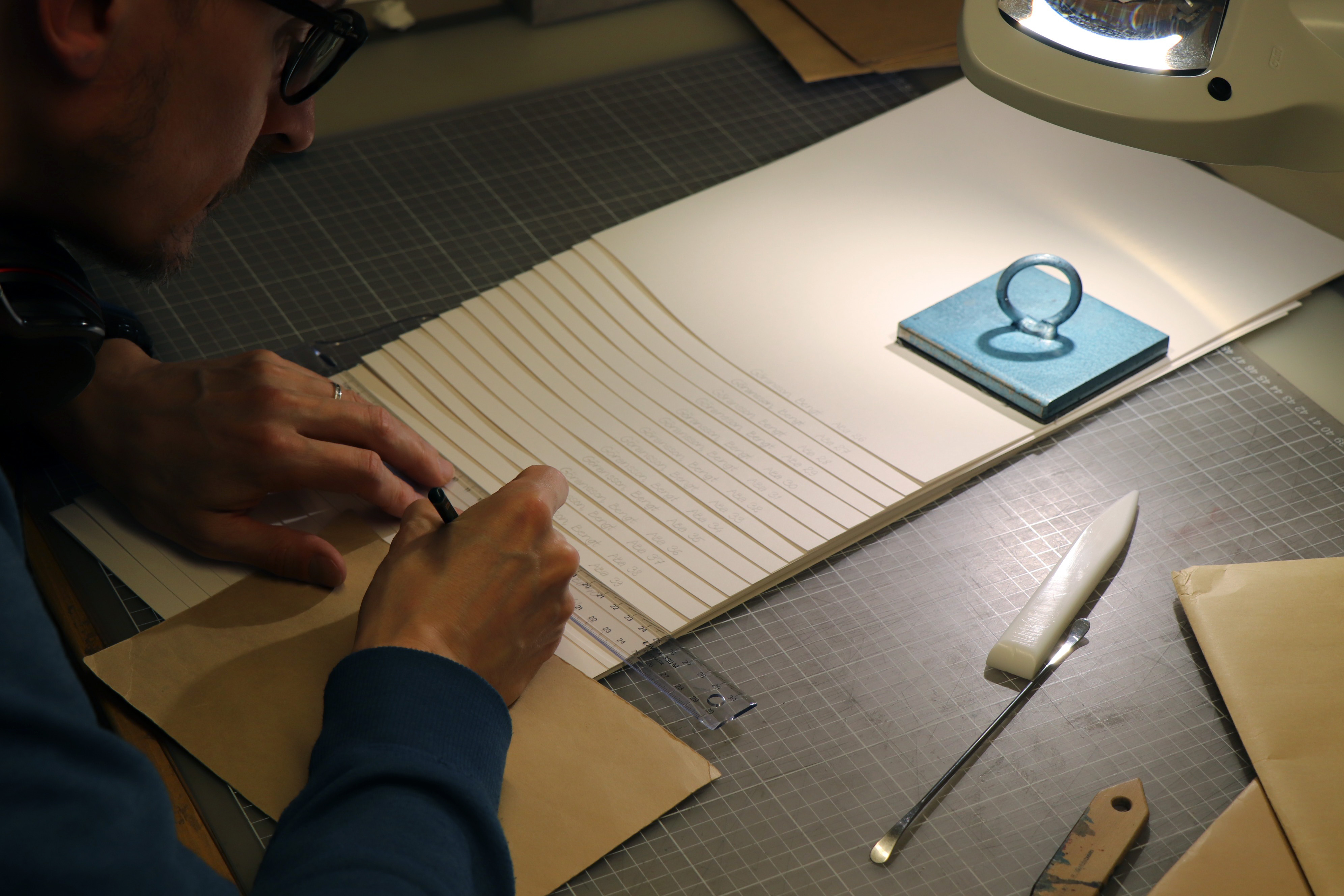 En man håller på att, med hjälp av en blyertspenna och en linjal, föra över text från ett kuvert till en bunt med nya, vita skyddsomslag som ligger på ett bord. På omslagen ligger en blå tyngd för att hålla dem på plats.