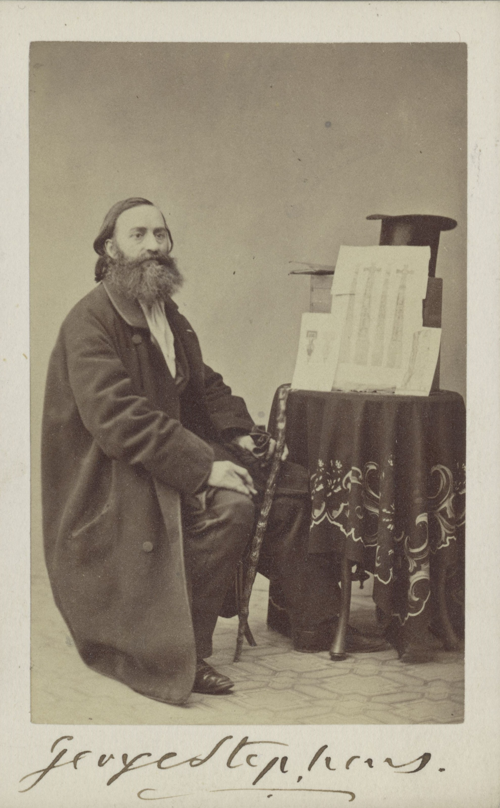 Fotografi av en långhårig man med långt skägg iförd en lång rock som sitter vid ett runt bord där flera teckningar är lutade mot en stapel som toppas av en upp och nervänd hatt. 