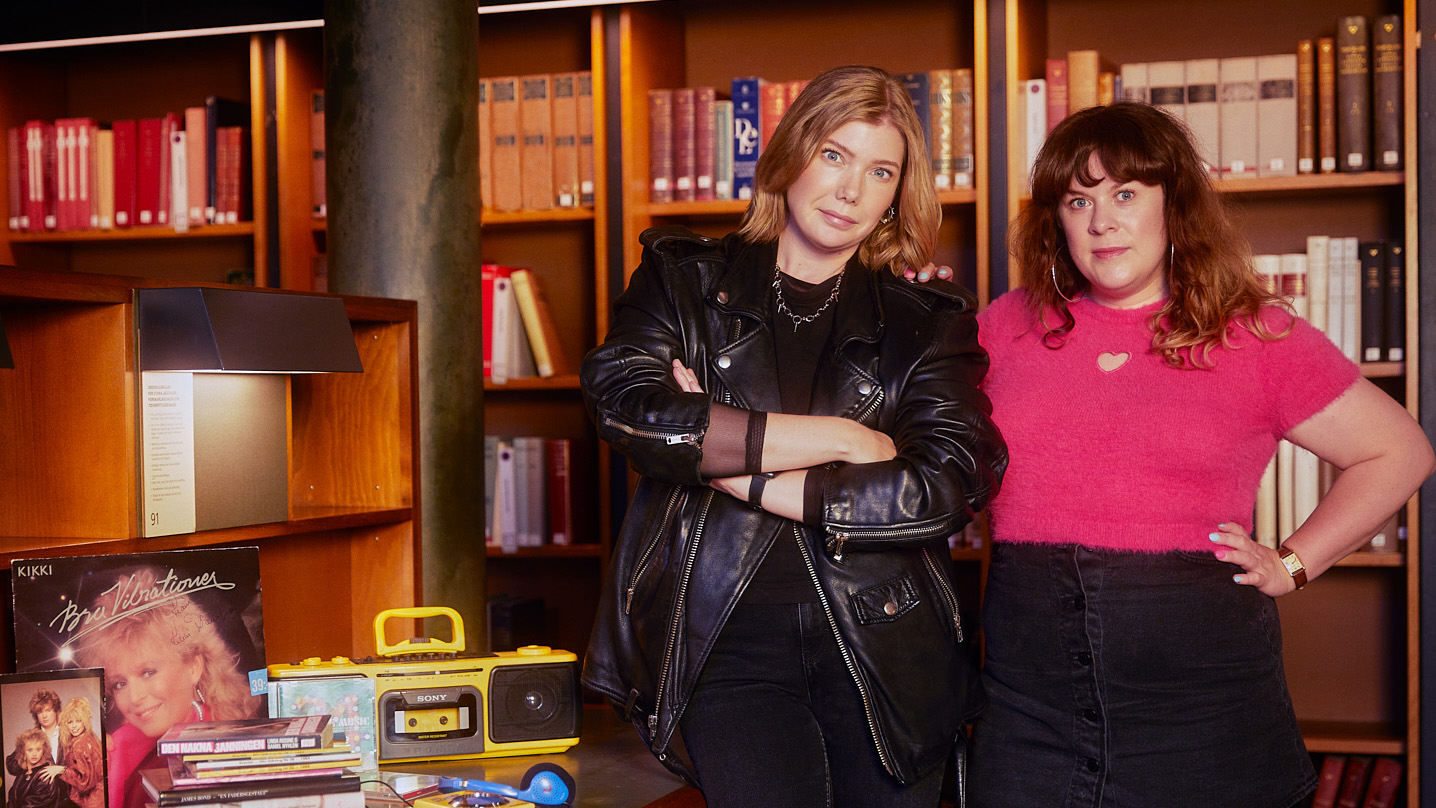 Två kvinnor står i ett bibliotek. På bordet bredvid dem syns en bandspelare, tidningar och en LP-skiva.