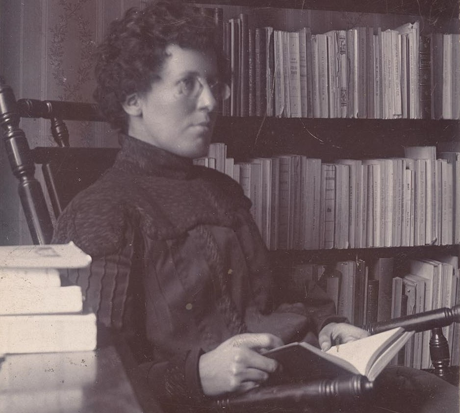 En ung kvinna med kort lockigt hår och glasögon sitter med en bok i knät i en gungstol framför en bokhylla. Hon är klädd i en mörk höghalsad klänning med långa ärmar.