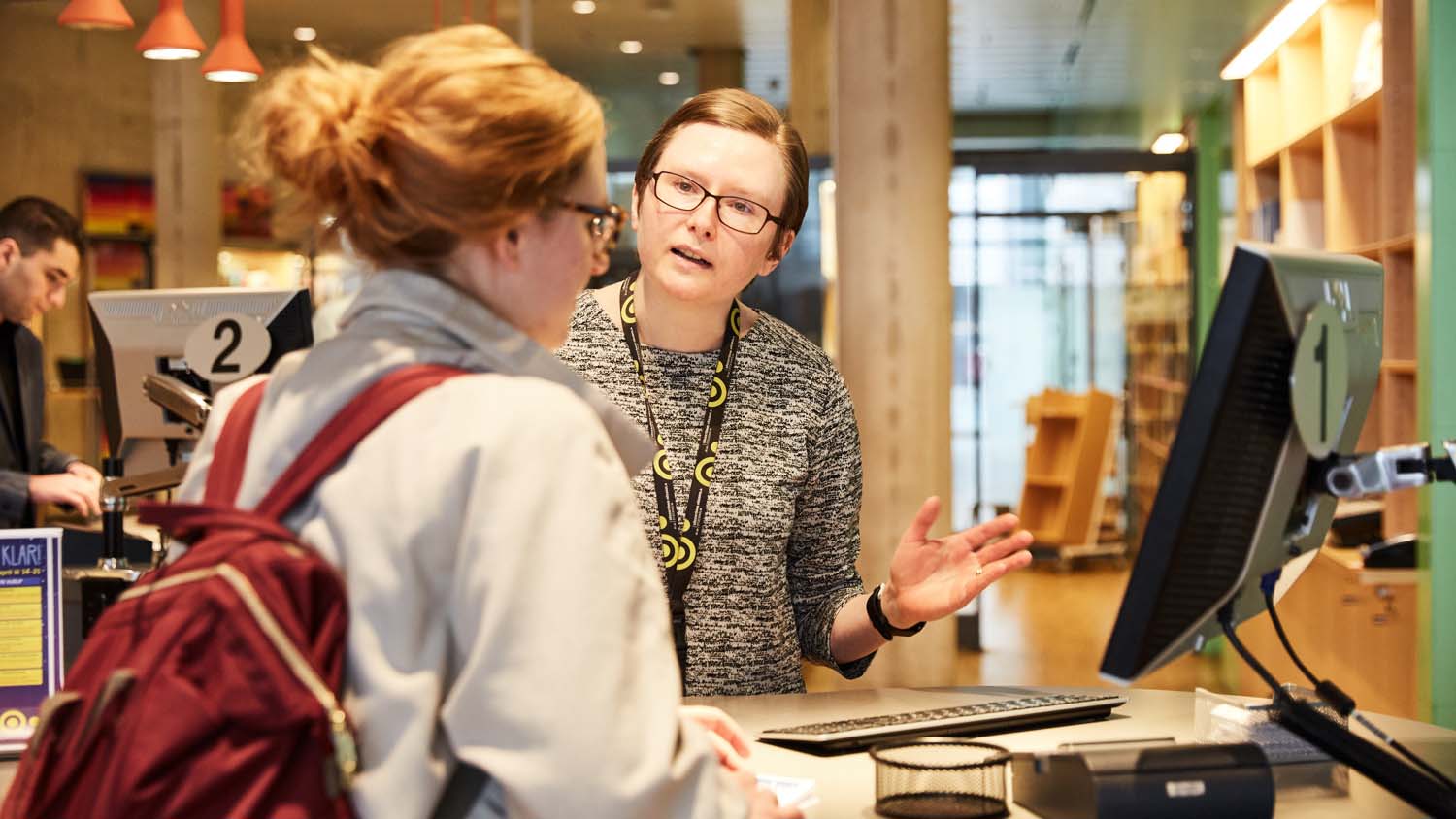 Två personer står vid en dator i en biblioteksmiljö.
