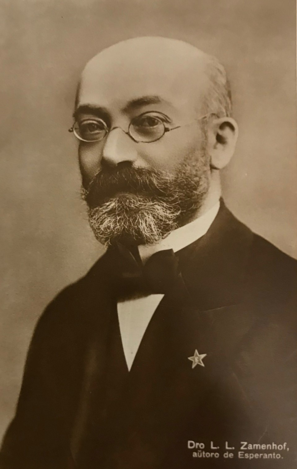 Porträtt av en skallig man med gråsprängt helskägg och ovala glasögon. På kavajslaget bär han en stjärna, en symbol för esperantospråket.