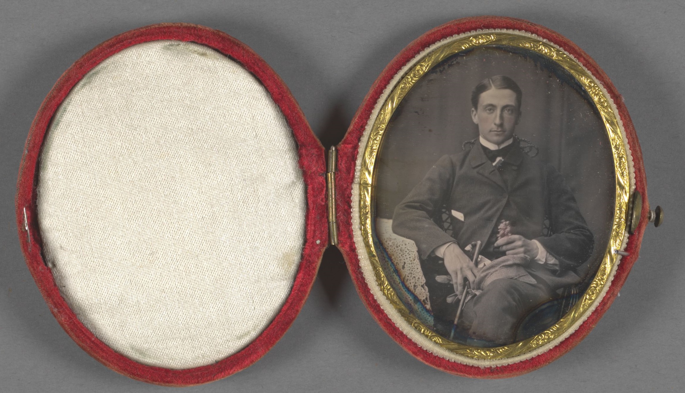 Greve C. G. Wrangel sitter tillbakalutad i rock och kravatt med korsade ben. I vänster hand håller han en käpp, handskar och en keps och i höger hand en blomma. Fotografiet är monterat i en oval ask.