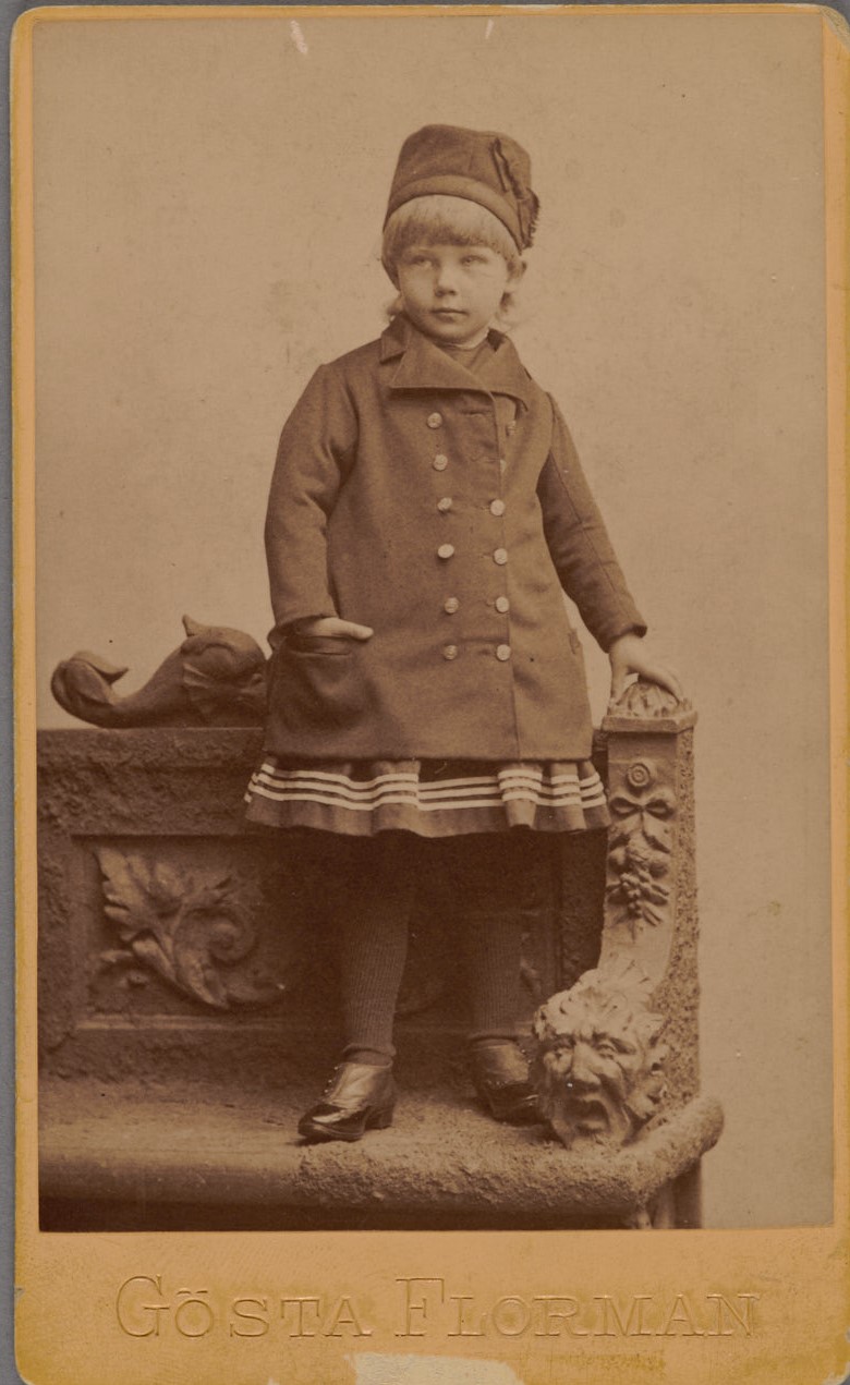 Fotografi av en liten flicka stående på en bänk iförd mössa och dubbelknäppt kappa
