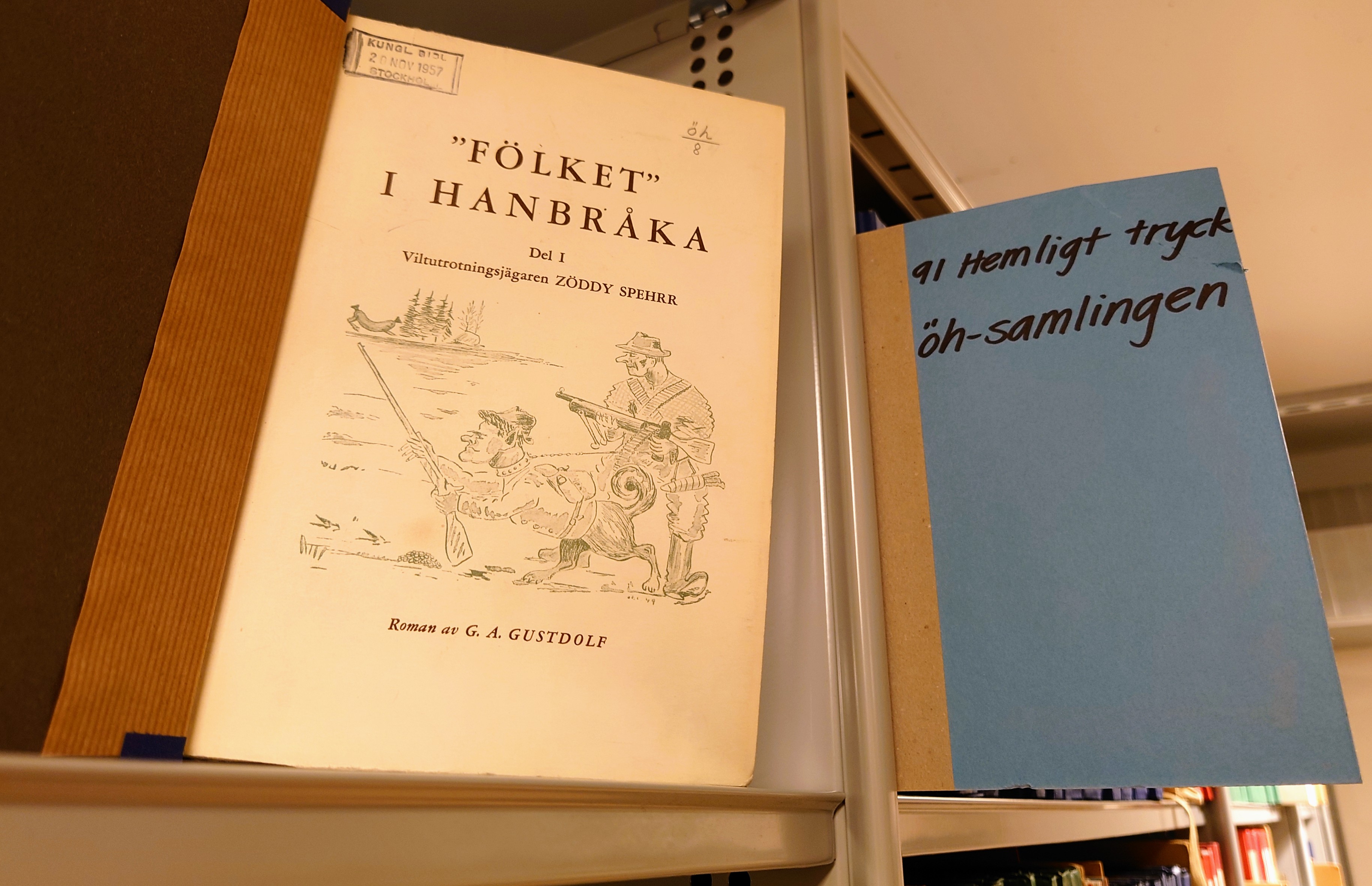 Uppslagen bok med titeln "Fölket" i Hanbråka på grå metallhylla bredvid blå skylt med texten 91 Hemligt tryck öh-samlingen.