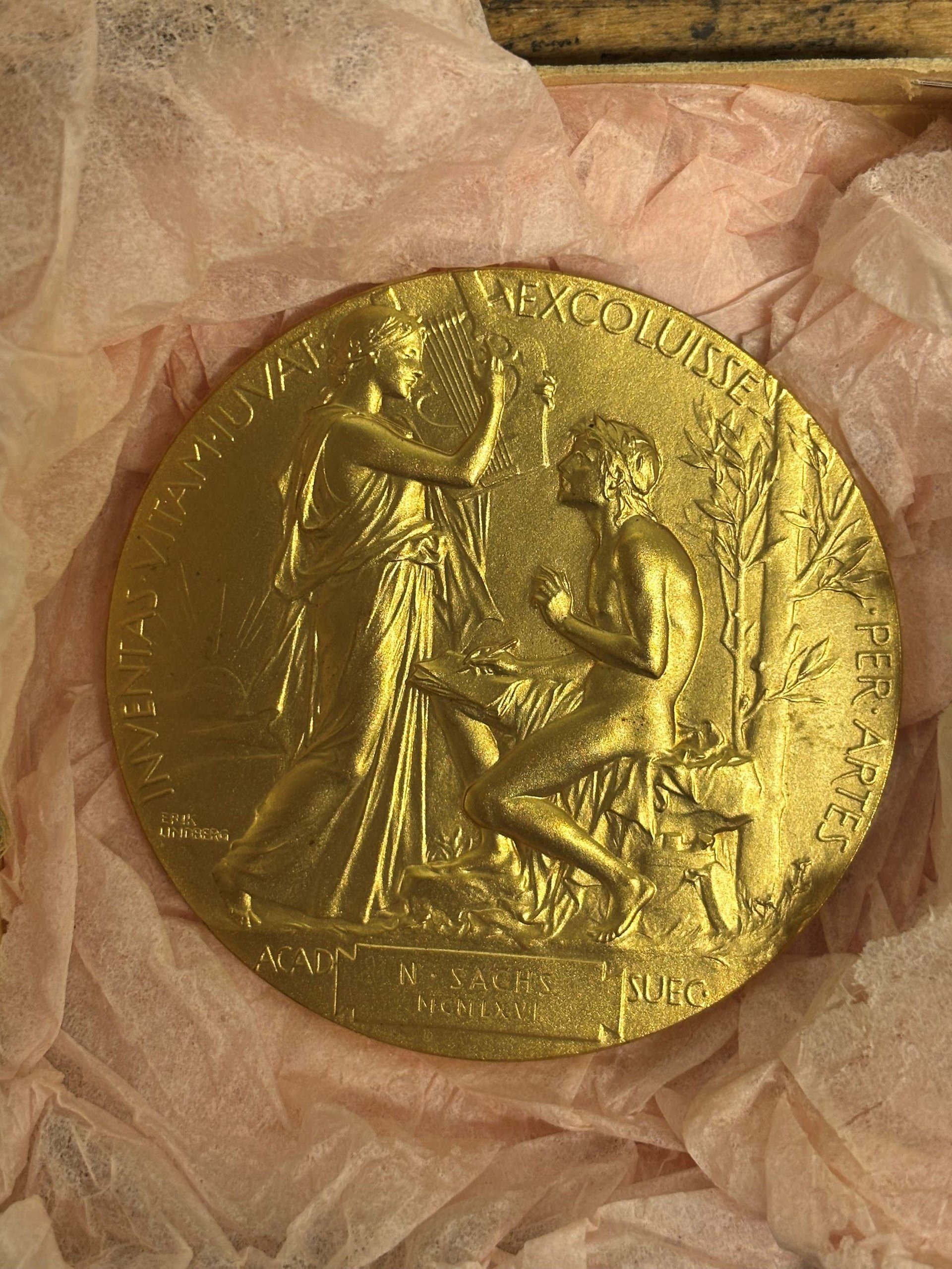 Guldfärgad medalj föreställande en stående kvinna och en sittande man i relief. Kvinnan håller upp en lyra, mannen håller i skrivdon. Runt medaljen löper en text i latin och i nedre delen syns namnet N. Sachs.