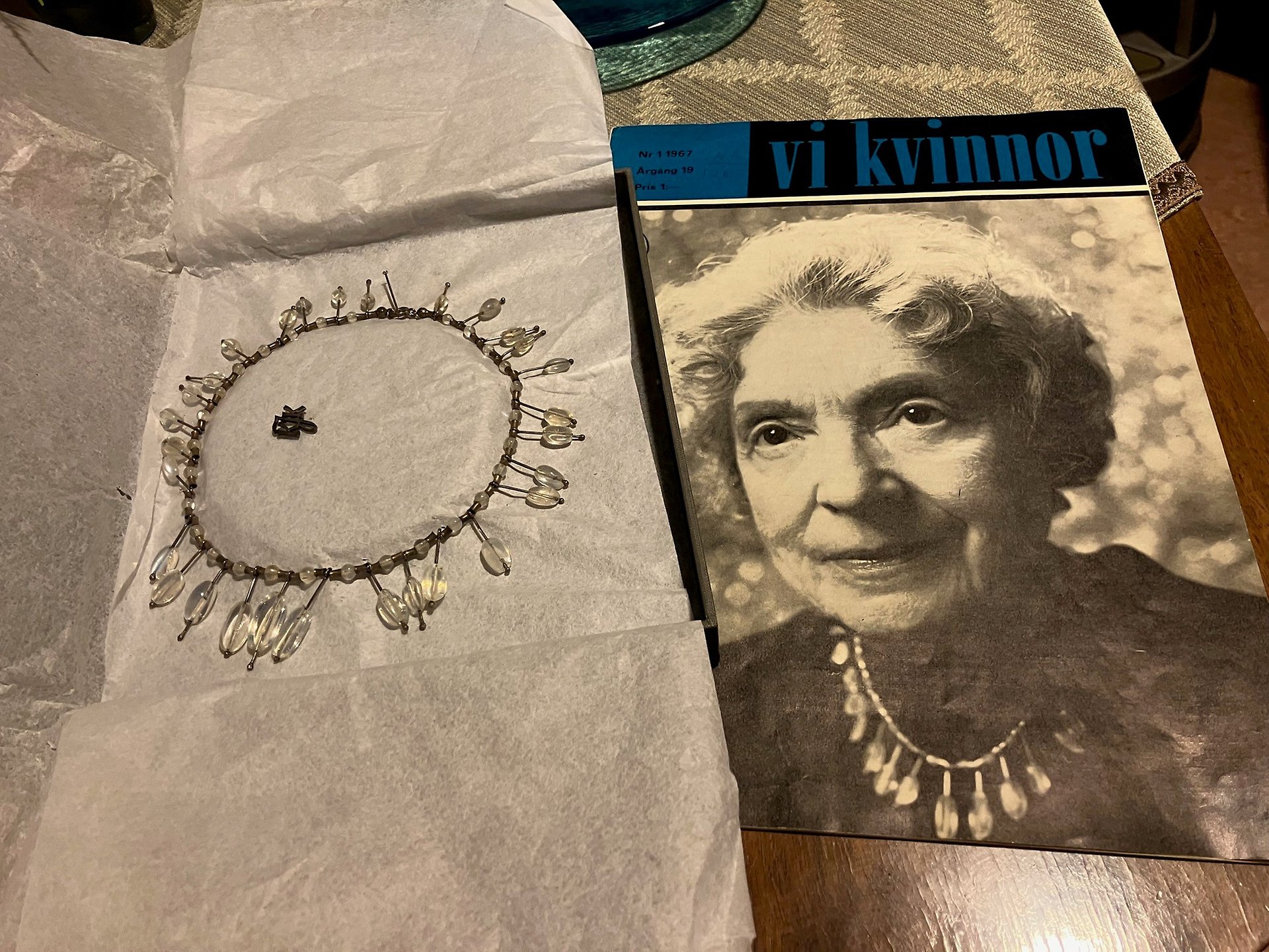Färgfoto med ett upplagt halsband prytt av genomskinliga stenar runtom. Bredvid syns framsidan på en tidskrift med ansiktet på en äldre dam som bär halsbandet.