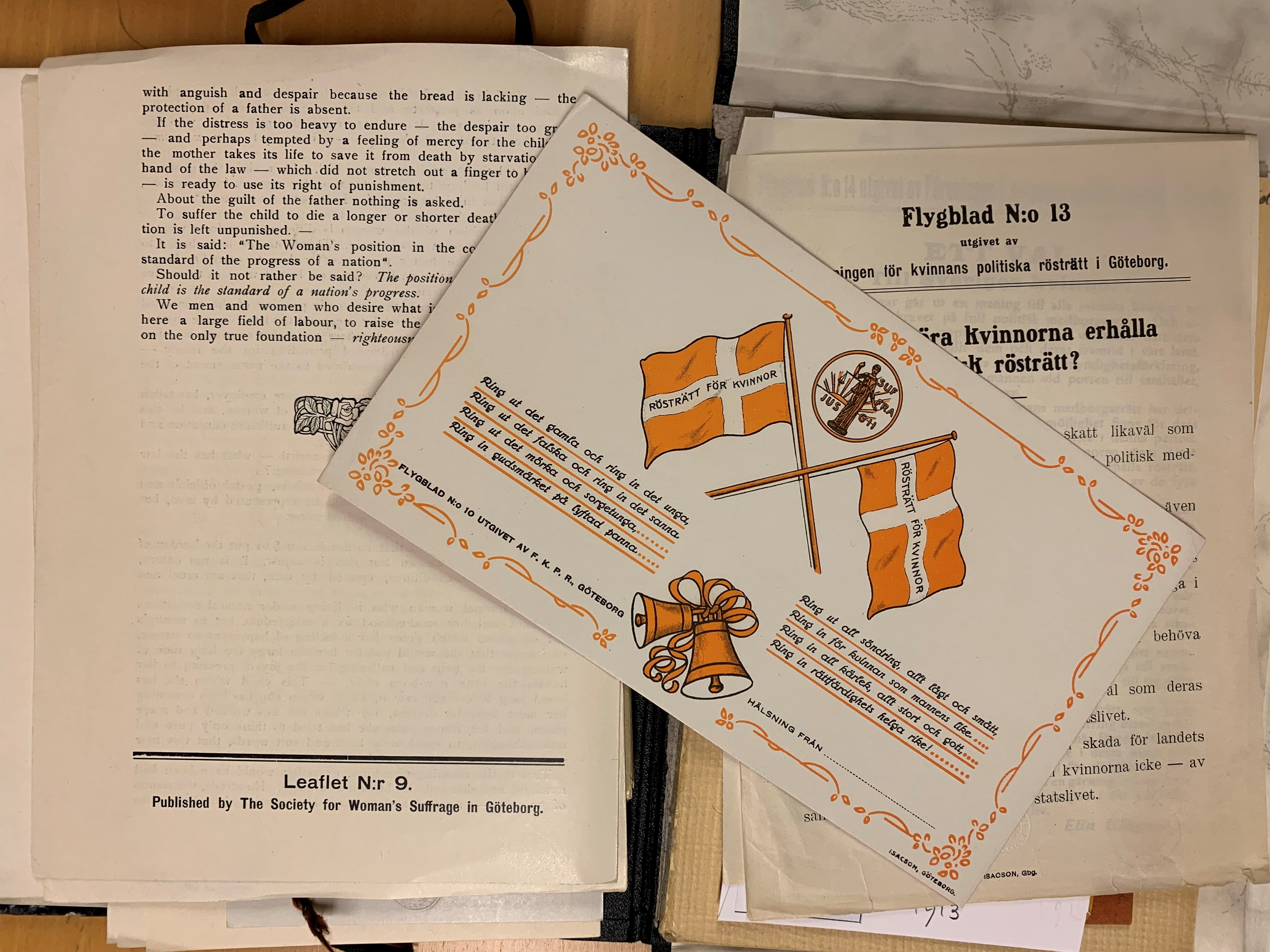 Två broschyrer med tryckt text, ovanpå ligger ett vykort med flaggor. 