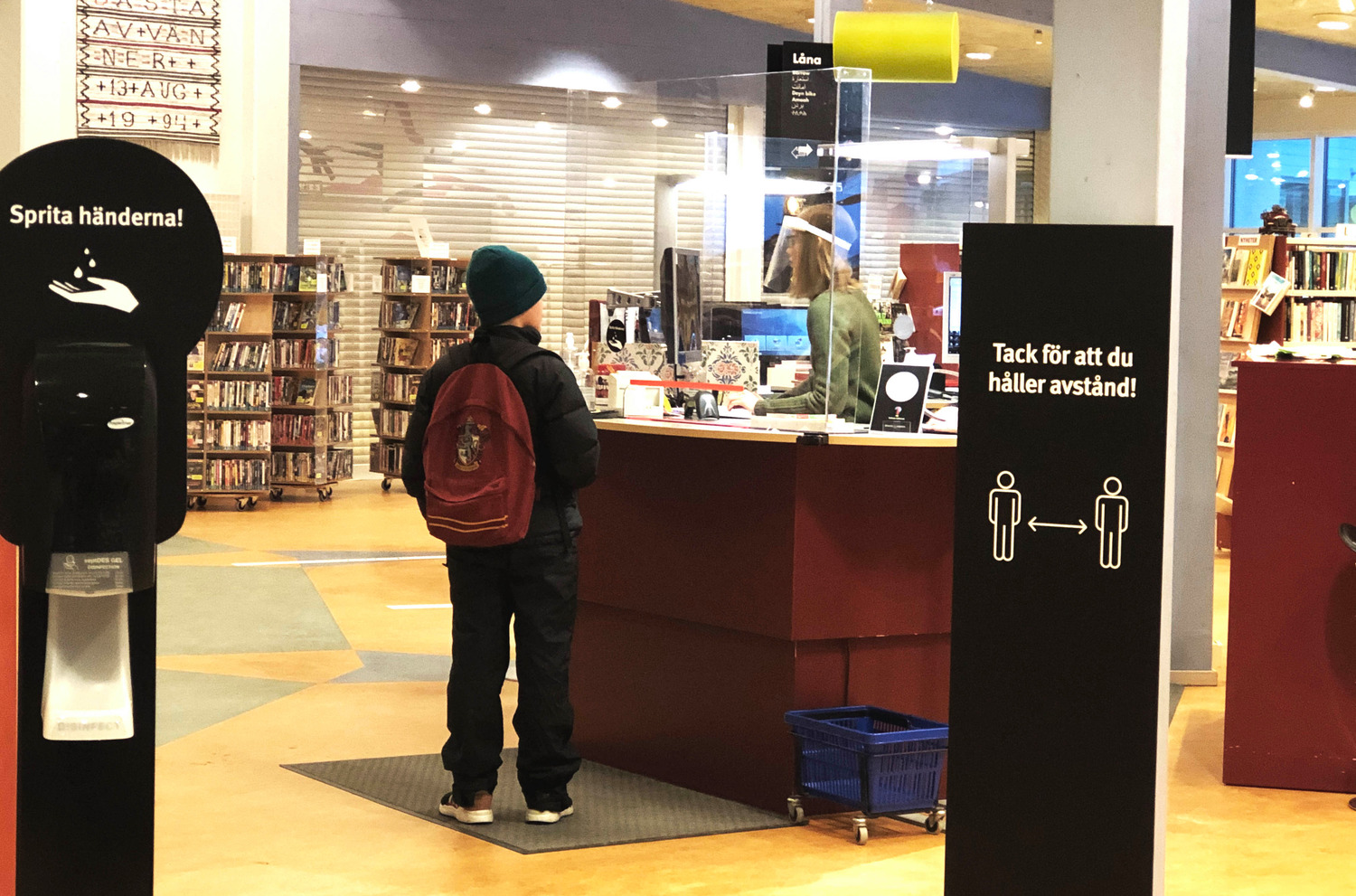 Ett barn står vid en biblioteksdisk, där personalen bär visir. Bakom barnet syns en behållare med handsprit och en skylt som uppmanar att hålla avstånd.