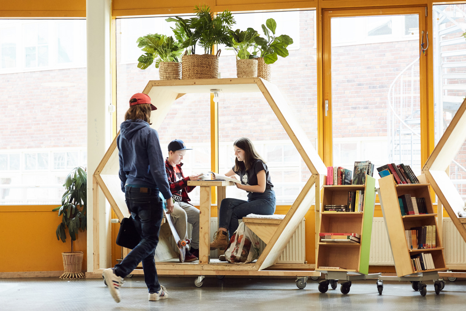 En person syns gående mot två personer sittandes i en hexagonformad studiehubb på högkant, i skolbibliotek, fryshuset.