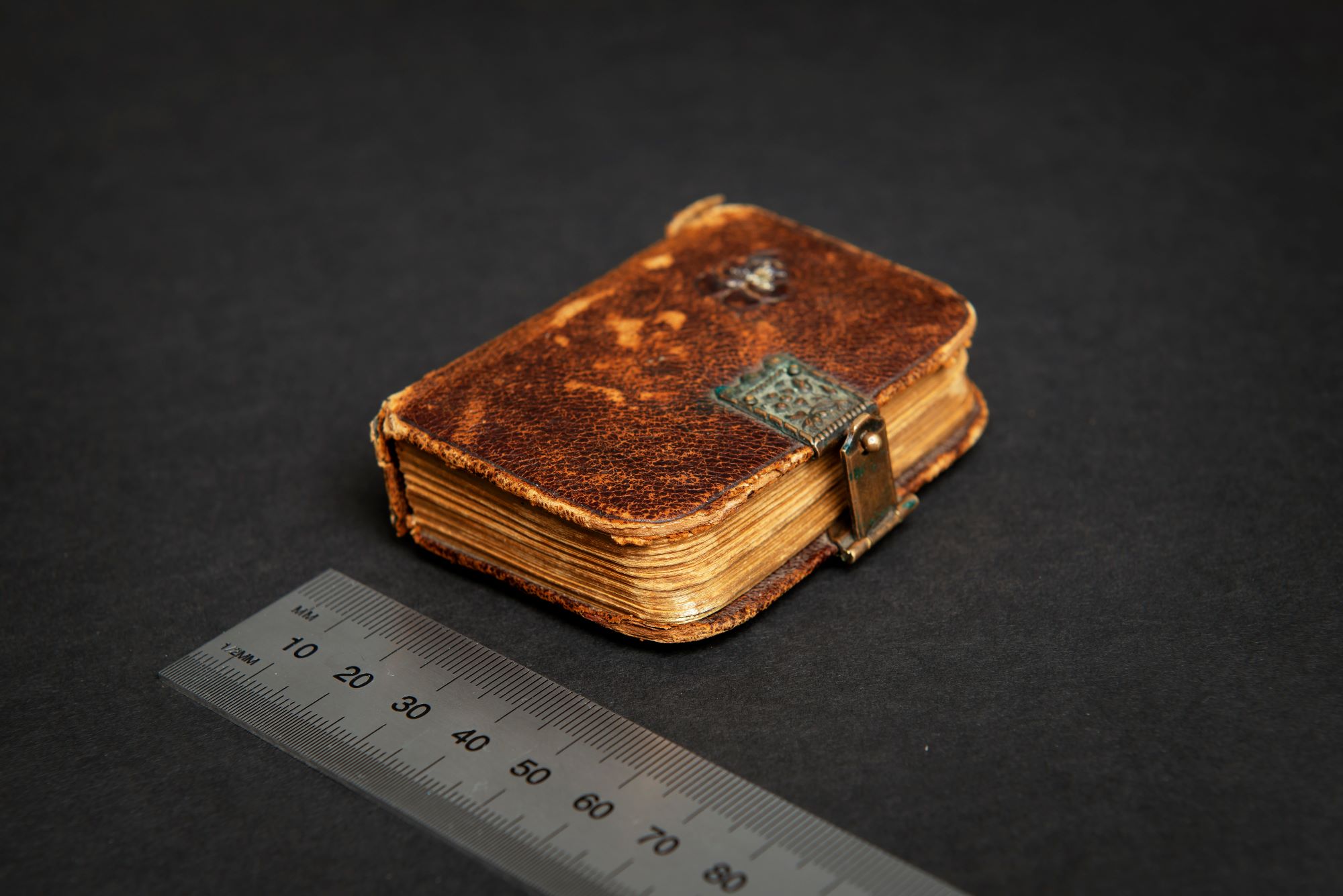 En liten anteckningsbok bunden i brunt läder med ett metallspänne ligger ovanför en linjal, som visar att boken är 5 centimeter bred.