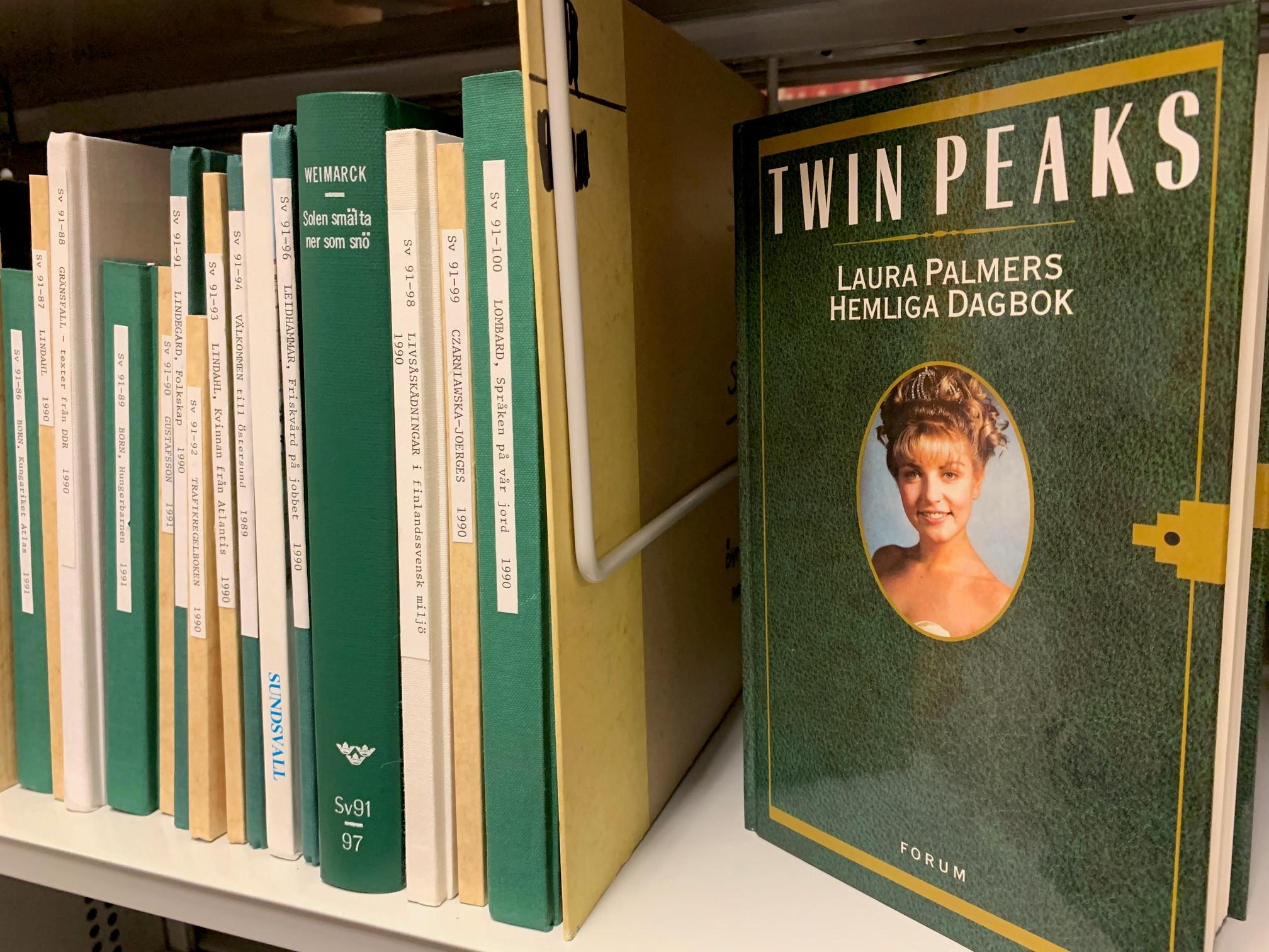 Bokhylla med tunna biblioteksbundna bokryggar, till höger står en bok med synligt omslag som föreställer en grön dagbok med ett ovalt porträtt av en ung flicka med diadem och uppsatt hår.