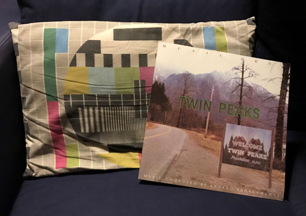 Ett skivomsalg till en vinylskiva med musiken till tv-serien Twin Peaks lutad mot en soffkudde med motiv som föreställer TV:s testbild