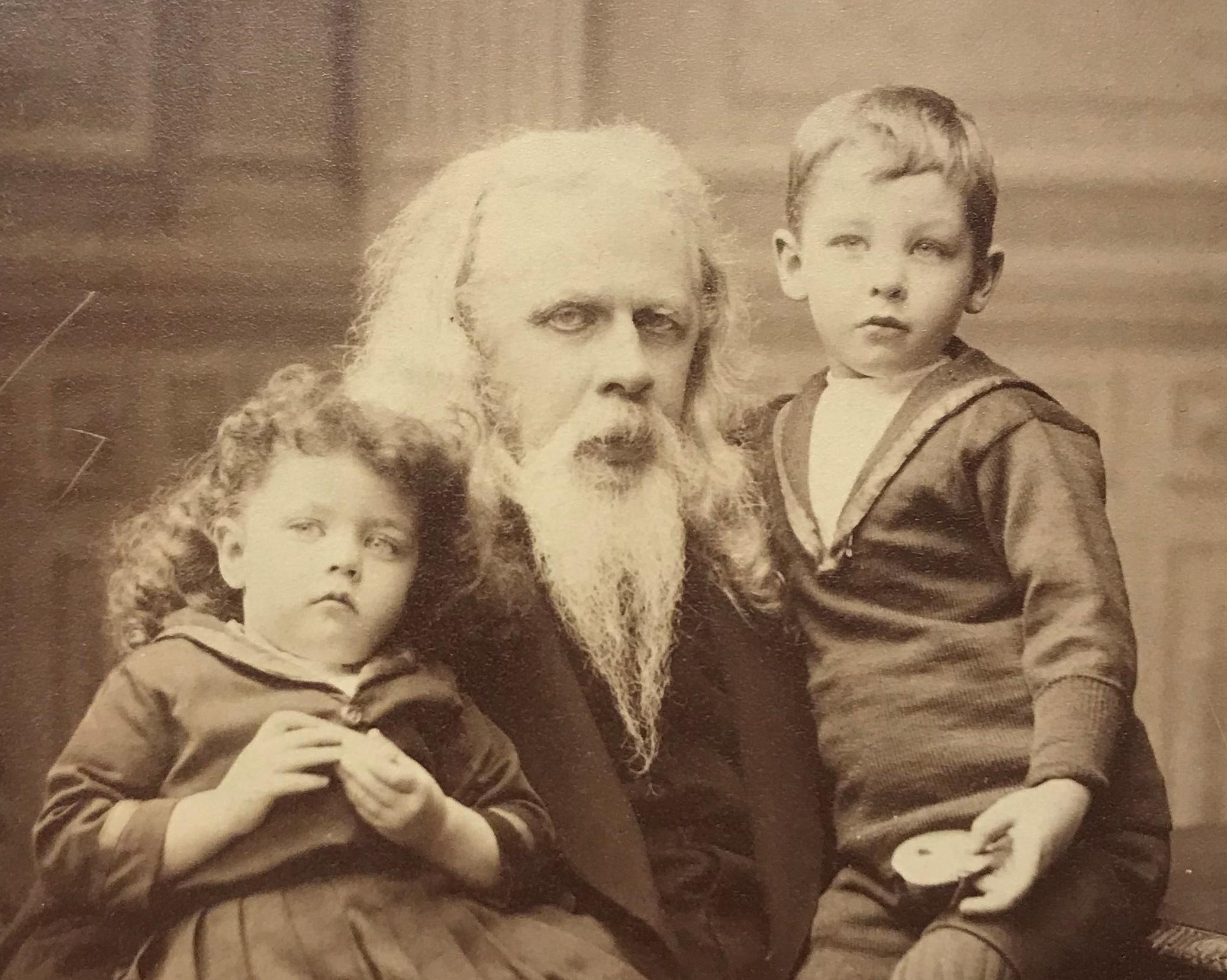 Fotografi av en långhårig man med långt vitt skägg som håller om två barn, en pojke och en flicka klädda i sjömanskostymer och snörkängor.