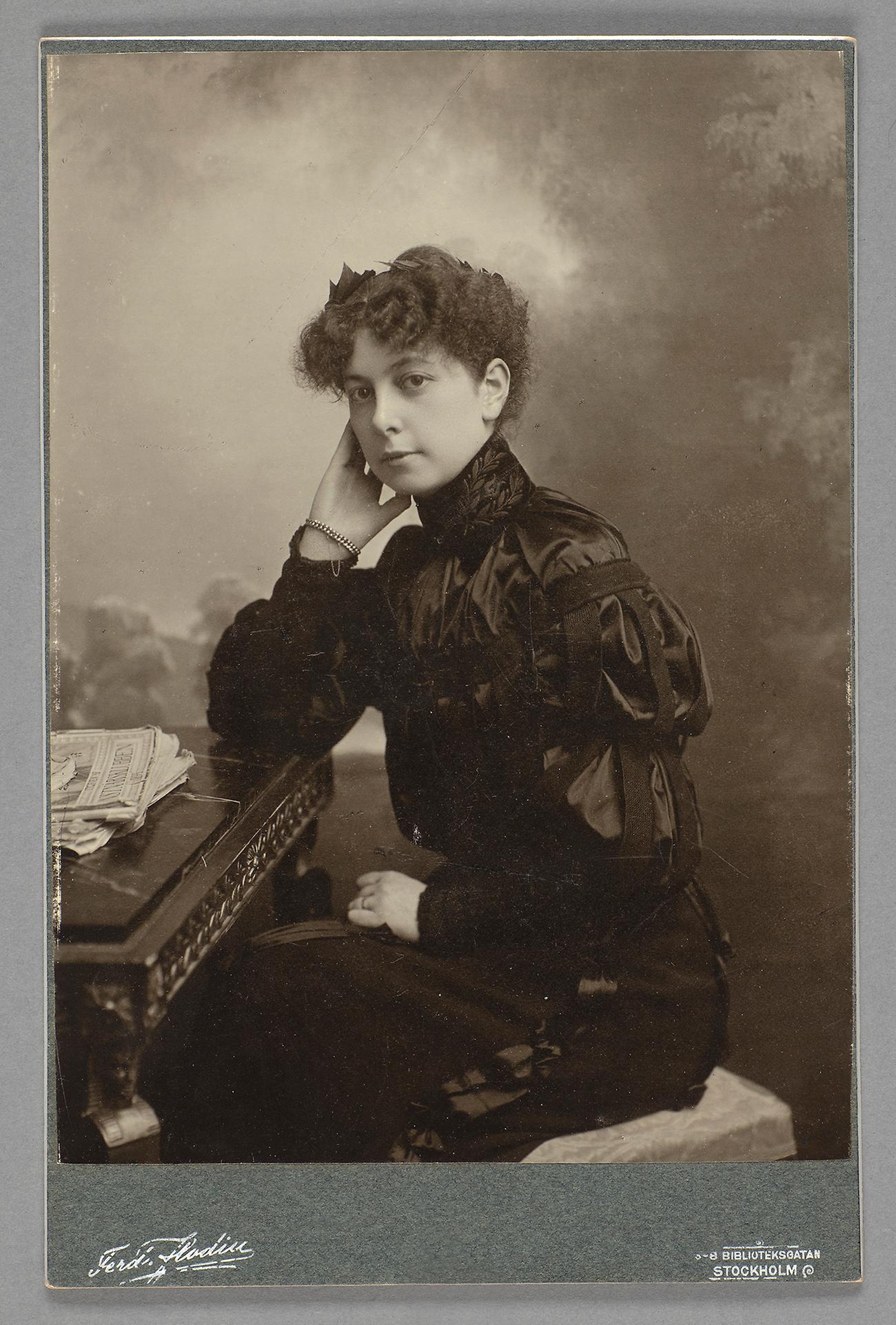Svartvitt fotografi med ung kvinna uppklädd i svarta kläder sittande på en stol vid ett bord