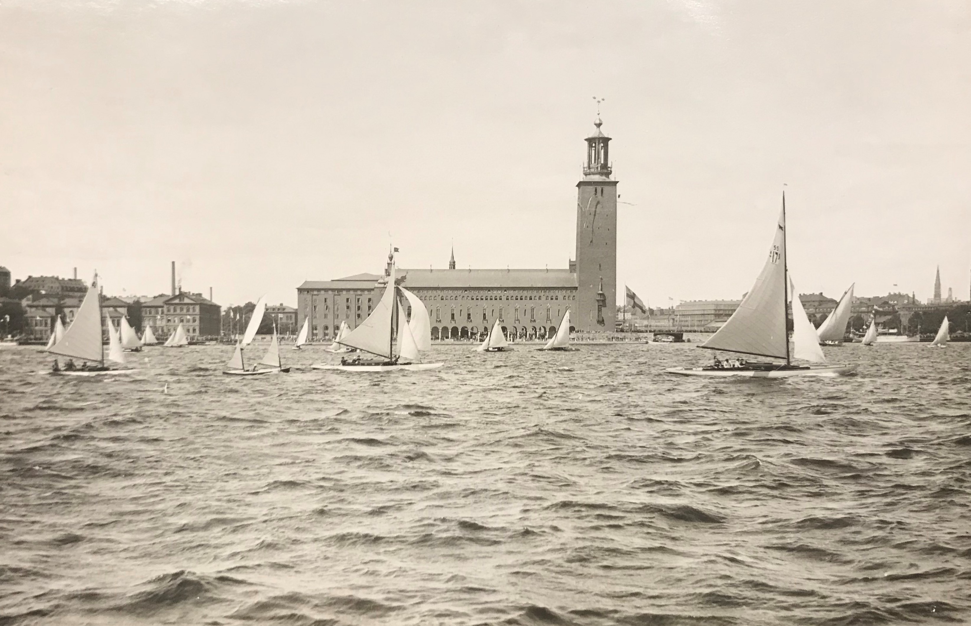 Svartvitt fotografi föreställande en mängd segelbåtar och roddbåtar i vattnet framför Stockholms stadshus.