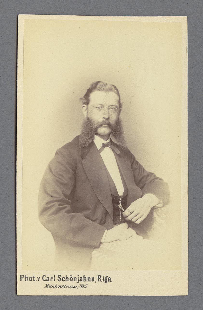 Porträtt av Axel Frisk sittande i en stol iklädd kavaj, kravatt och glasögon.