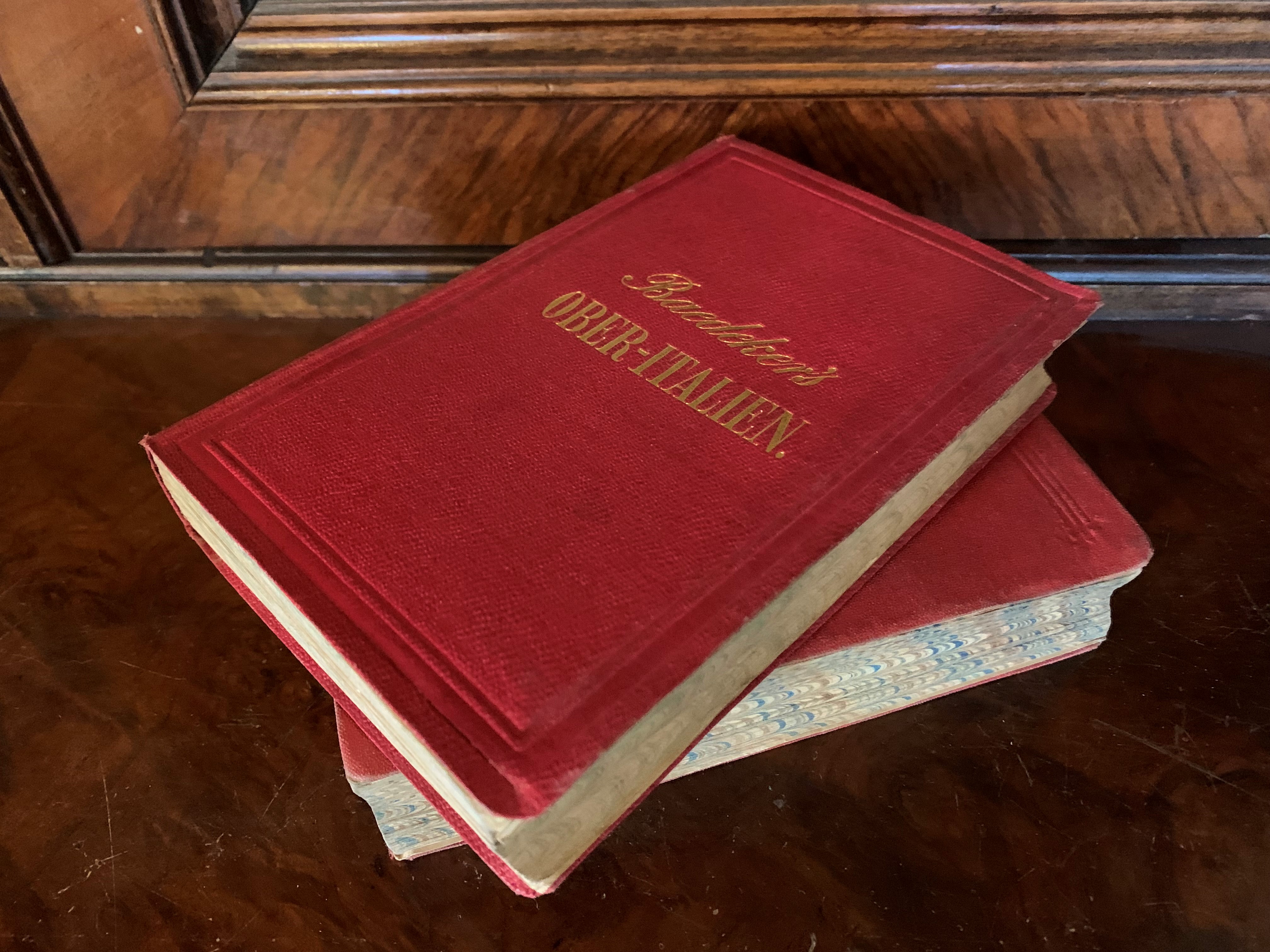 Två guideböcker ur med röda pärmar från Baedekers förlag liggande ovanpå en möbel i mörkt trä.