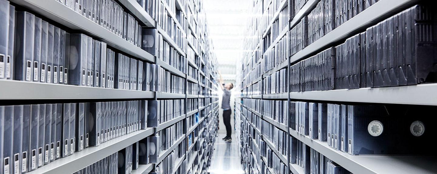 A room with floor-to-ceiling shelves filled with VHS tapes. Someone stretches up to take out a tape.