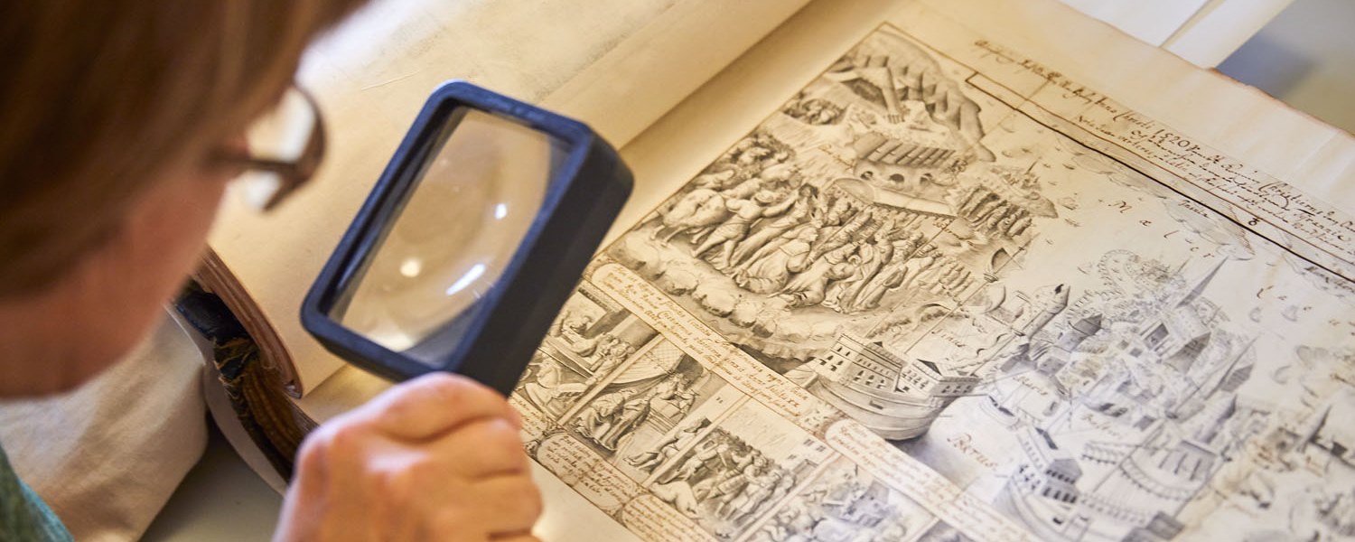 Någon tittar på handmålade bilder i en gammal bok genom ett förstoringsglas.