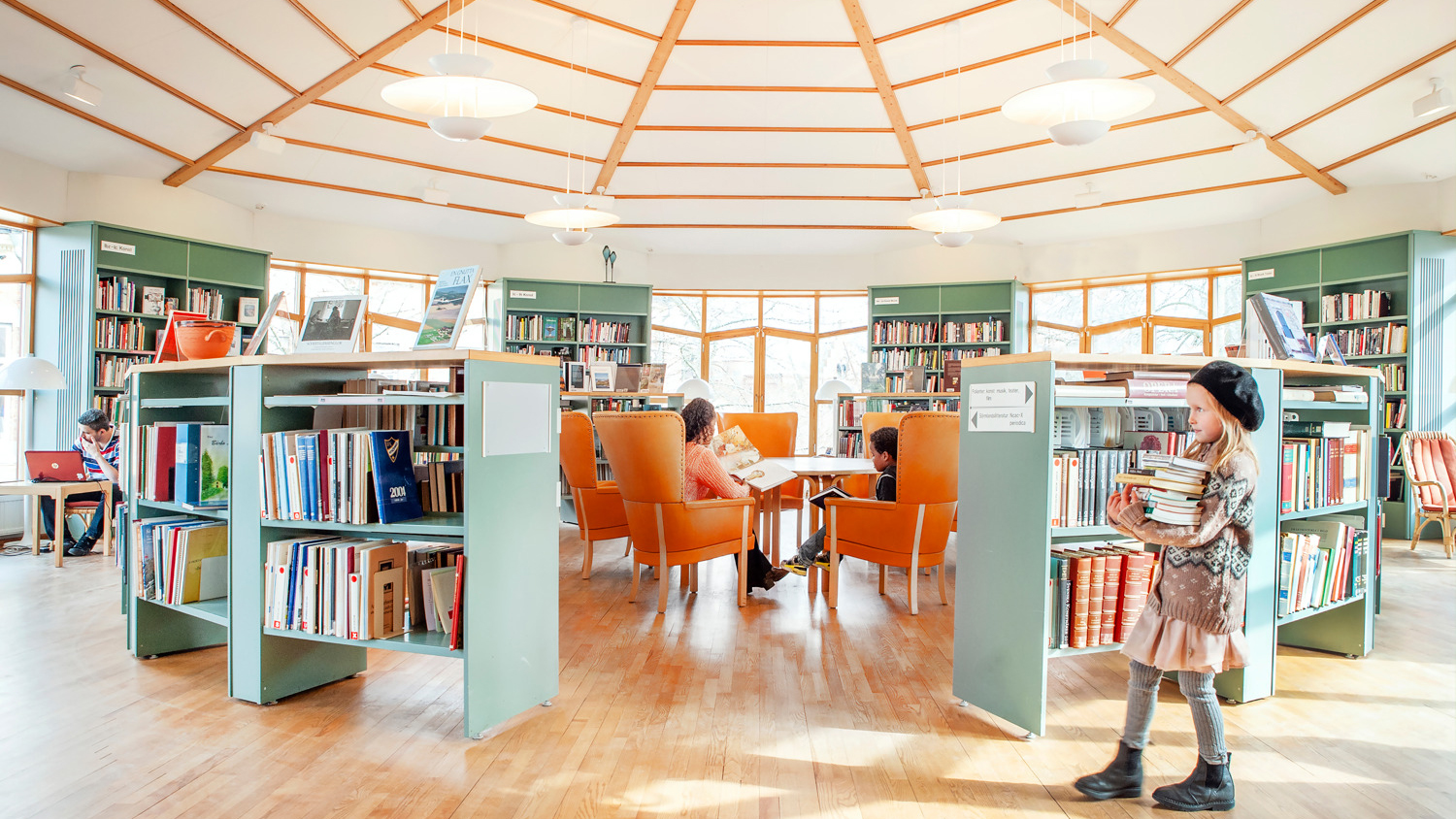 En flicka i basker bär böcker i en öppen, ljus bibliotekslokal.