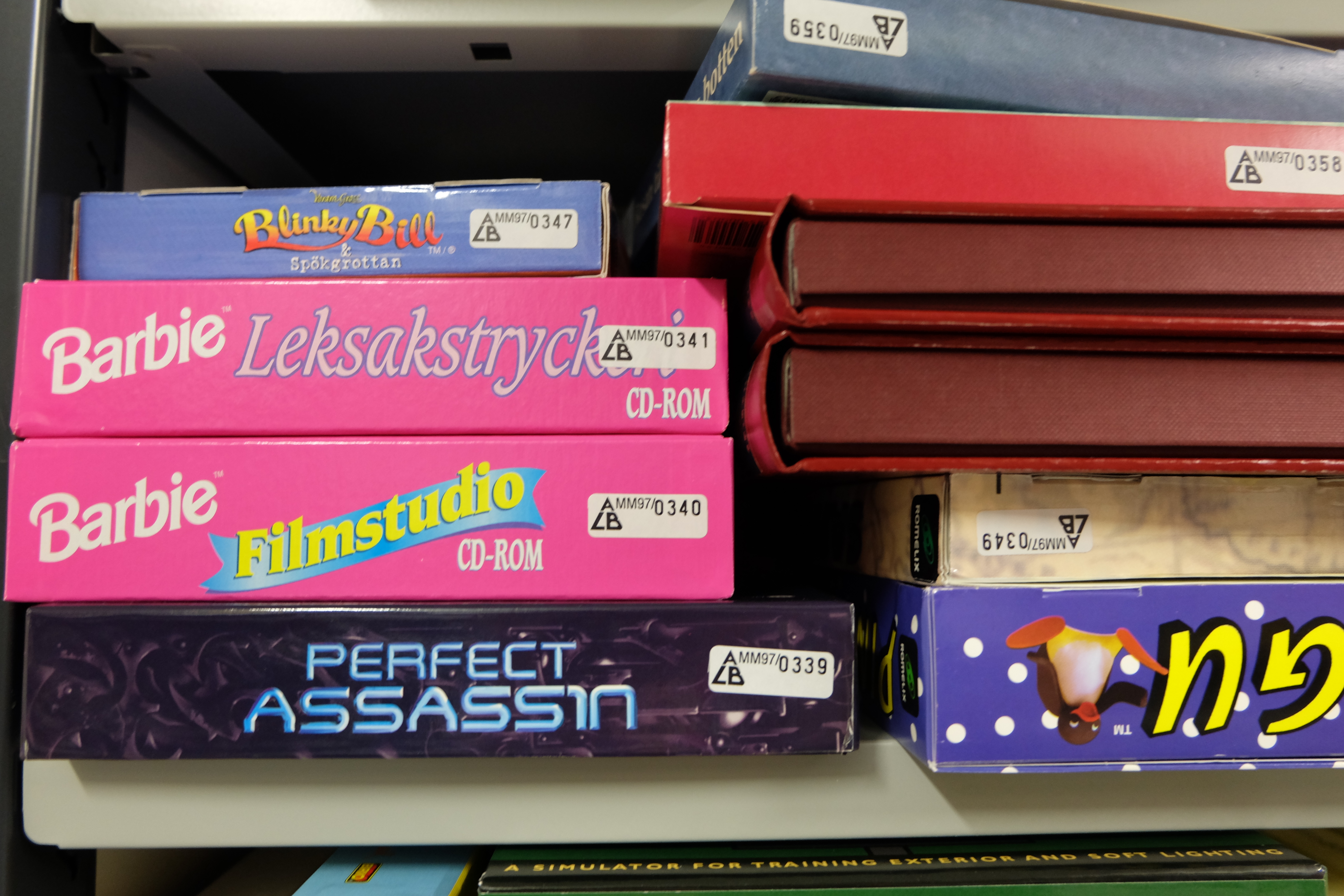 Hylla med datorspel. Längst fram syns några rosa omslag med texten "Barbie".