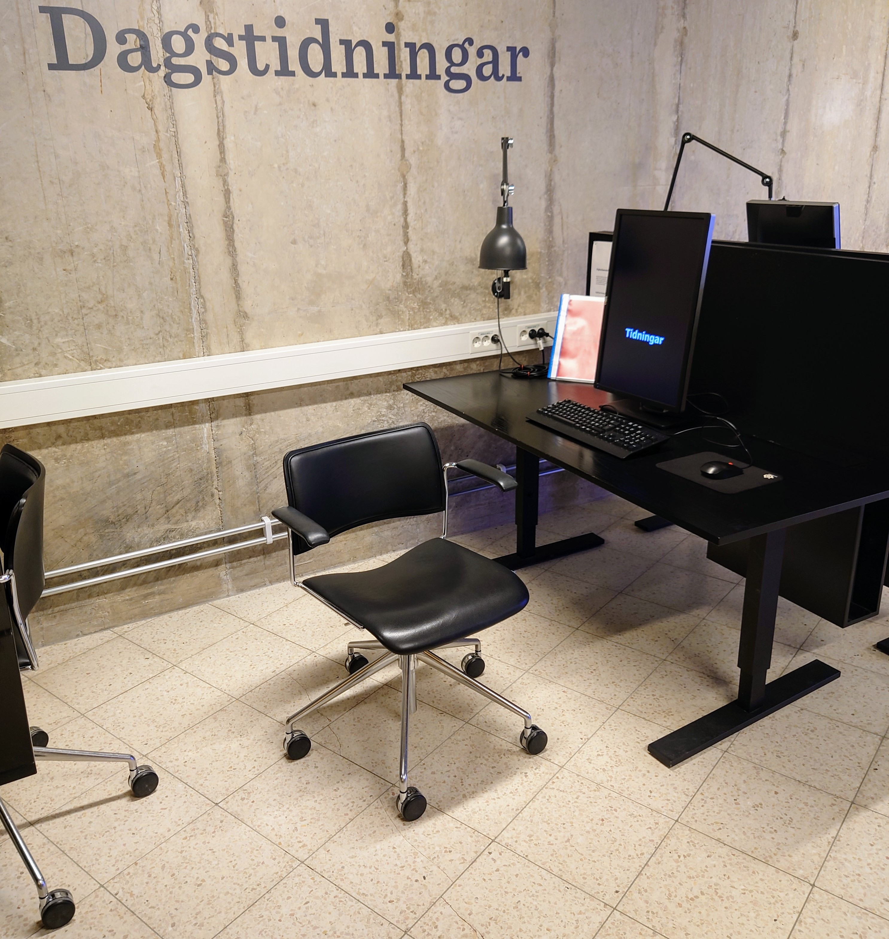 Fotografi på skrivbord där en dator med stor skärm står, på väggen texten Dagstidningar.