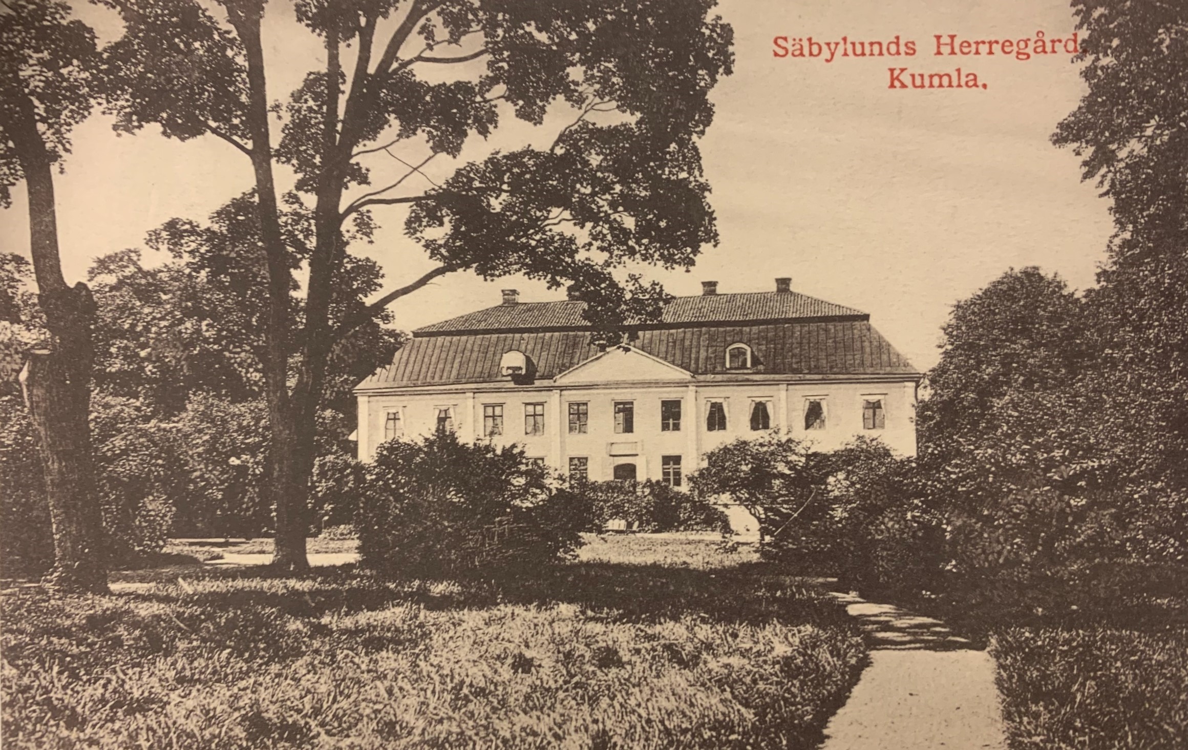 Svartvitt foto föreställande en herrgård i vitt trä med grönskande träd runt om.