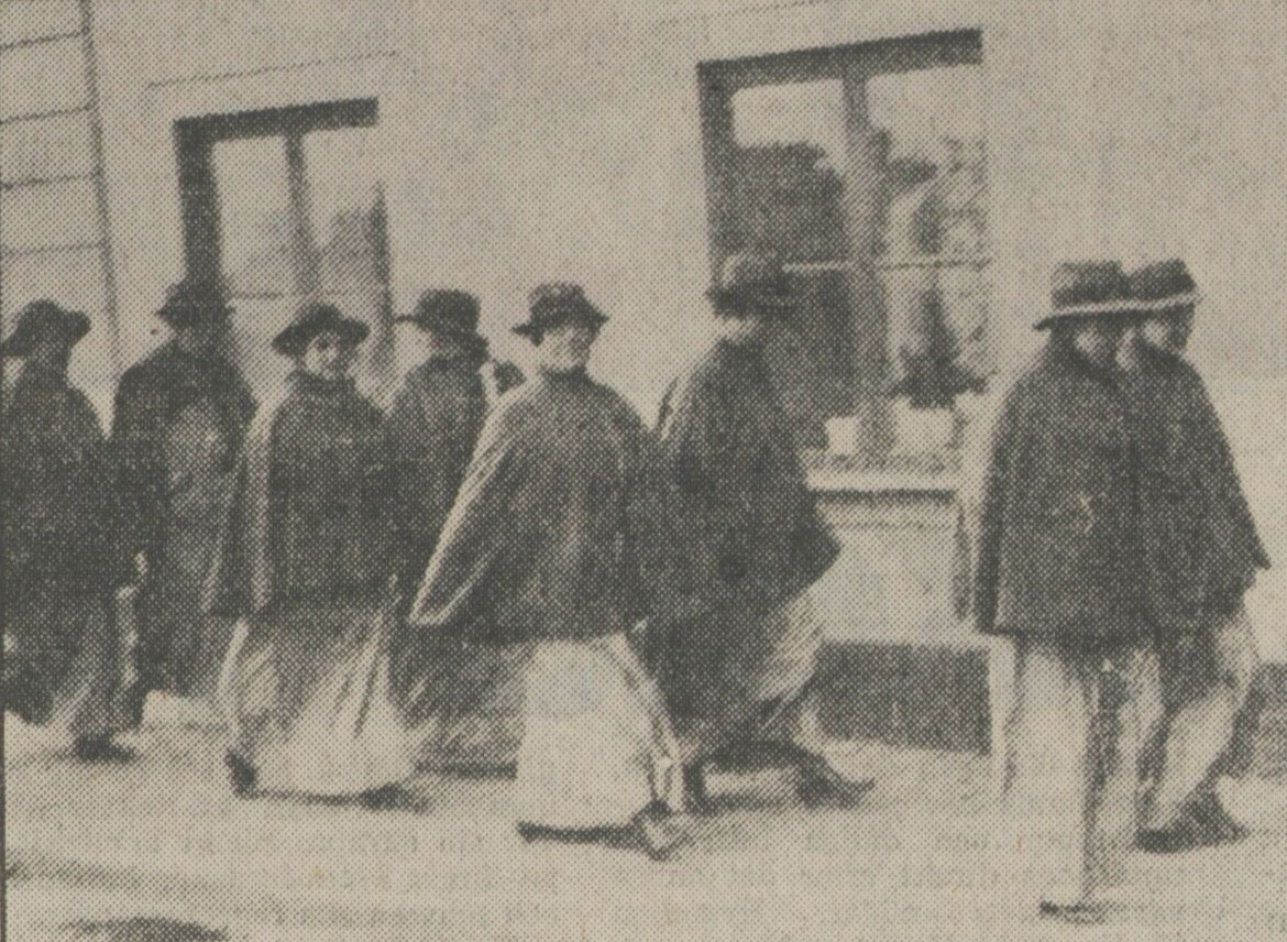 Grynigt och svartvitt fotografi av kvinnor med hattar, kappor och kjolar som kommer gående.