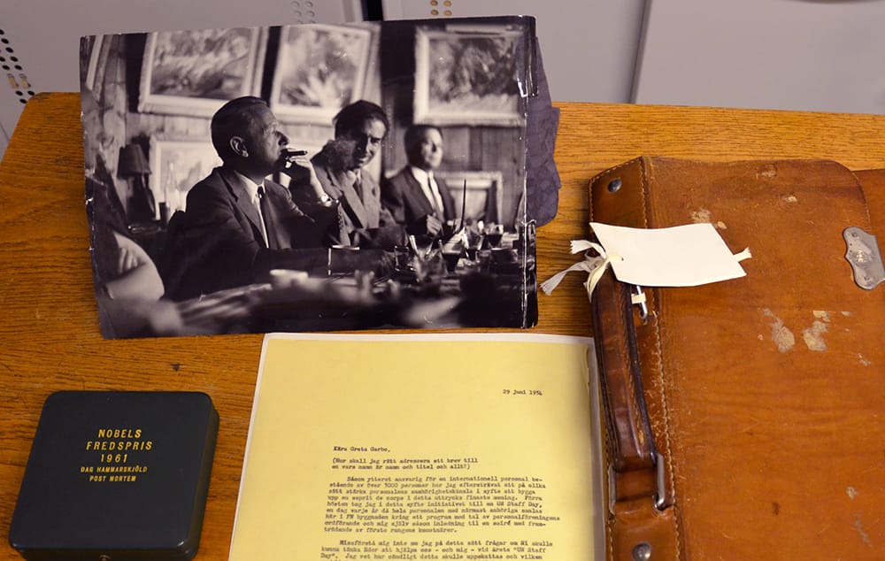 Ett svart fodral med Hammarskjölds nobelpris, ett svartvitt fotografi av honom rökandes cigarr, ett gult dokument och hans bruna portfölj.