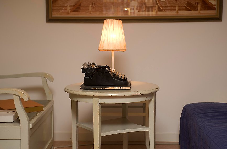 En skrivmaskin och en lampa står på ett litet runt bord. Till vänster syns en kökssoffa och till höger ett hörn av en säng. 