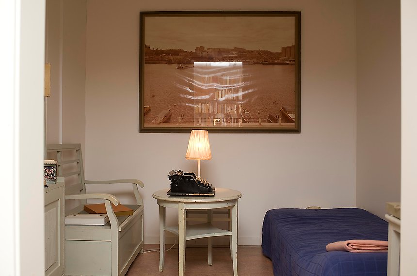 Ett rum med en kökssoffa till vänster, ett litet runt bord med en skrivmaskin och en lampa i mitten och en säng till höger.