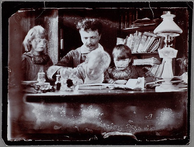 Svartvitt fotografi av en man med yvigt hår och tre barn vid ett skrivbord.