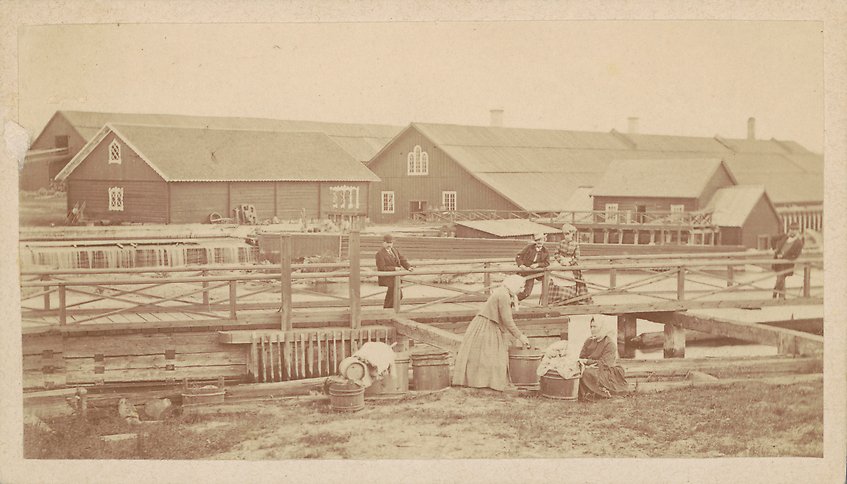 Svartvitt fotografi av två kvinnor i huckle som tvättar kläder framför en stor gårdsbyggnad.