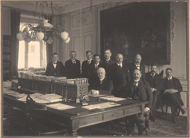 Svartvitt fotografi av en grupp män i kostymer bakom ett stort skrivbord.