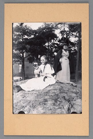 Svartvitt fotografi av en flicka som sitter på en bergskulle med en hund i famnen. Bakom hennes står en finklädd kvinna.
