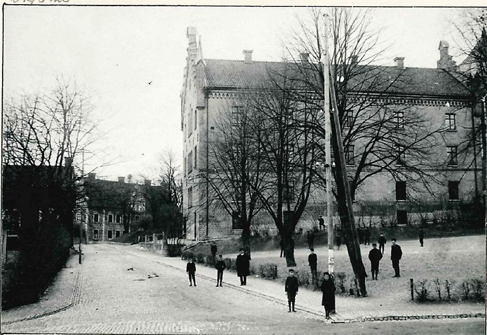 Äldre svartvitt fotografi av gathörn där barn och bara lövträd syns i förgrunden. I bakgrund stor tegelbyggnad.
