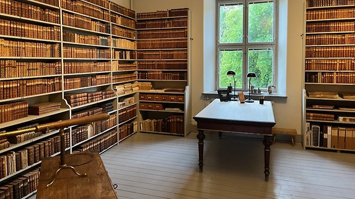 Ett skrivbord, gröna läslampor, en gammal kikare och bokhyllor med äldre böcker.