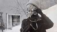 En person i vinterkläder står utomhus med en gammal kamera framför ansiktet.