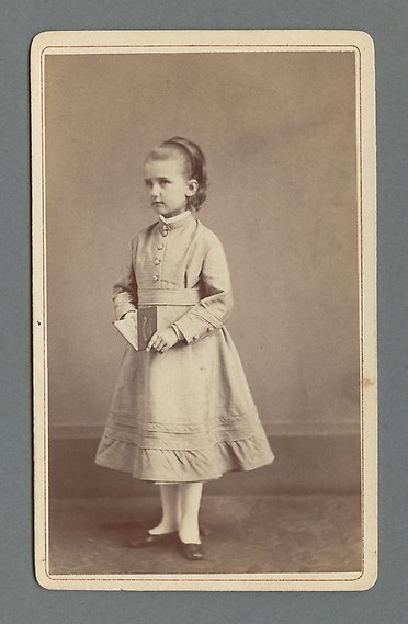 Svartvitt fotografi av ung flicka i vit klänning. I handen håller hon en uppslagen bok. 