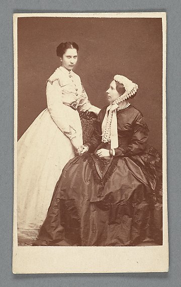 Svartvitt fotografi av en stående kvinna i vit klänning som håller en något äldre sittande kvinna i svart klänning i handen. 