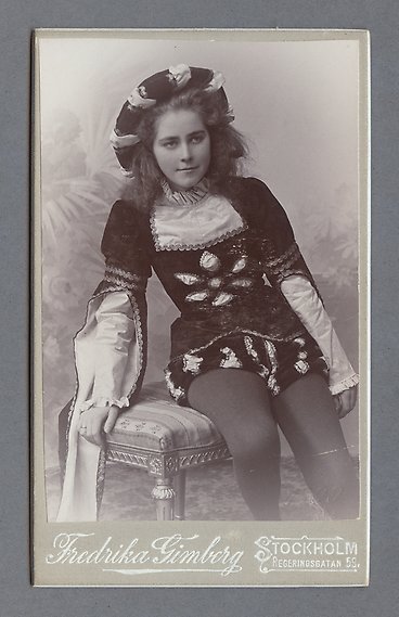 Svartvitt fotografi av kvinna i festlig klädnad. Hon sitter på en påkostad stoppad pall.