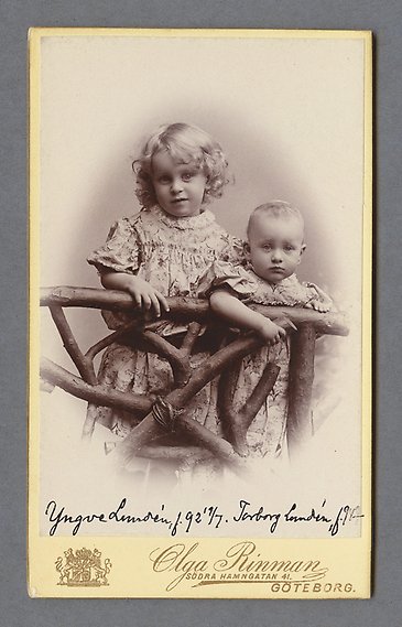Svartvitt fotografi av två små barn i blommiga klänningar som lutar sig mot ett gammalt trästaket.