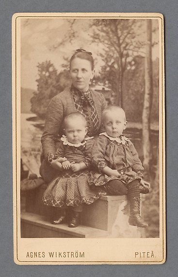 Svartvitt fotografi av kvinna med två små barn i matchande kläder i famnen. I bakgrunden en kuliss i naturstil.