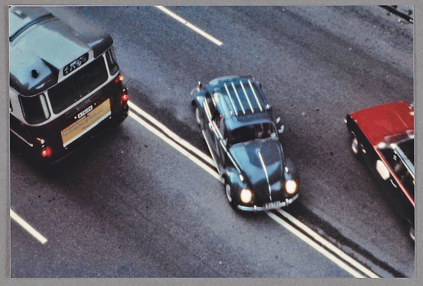 Färgfotografi taget snett uppifrån där en bil syns korsa heldragen linje i trafiken. Delar av en buss och en röd personbil syns också.
