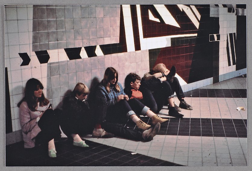 Färgfotografi där ungdomar syns sitta på golvet i en gång i tunnelbanan.